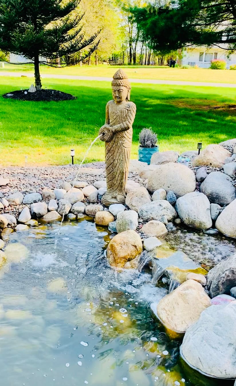Buddha statue fountain pouring into a boulder-edged pond on a green lawn