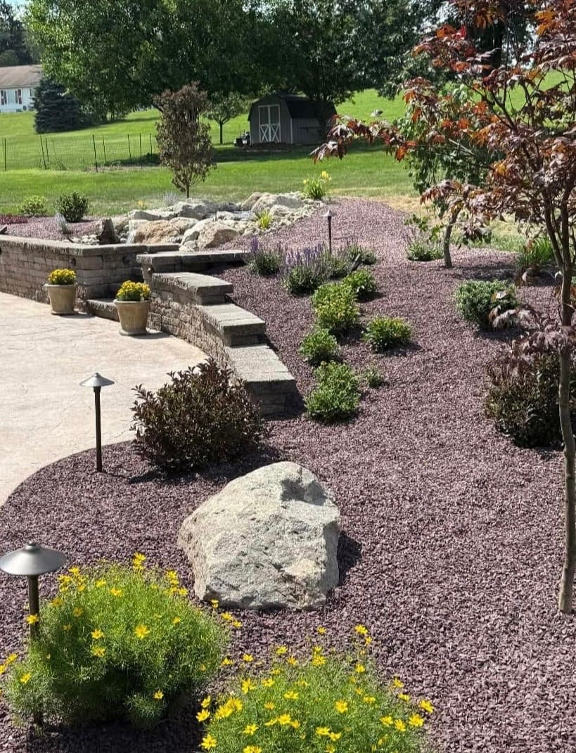 Curved block retaining wall with steps, gravel beds, and new plantings around a pondless feature